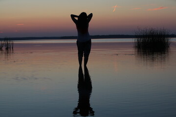 Silhouette of a girl standing in the water at sunset stretching with raised hands to her head