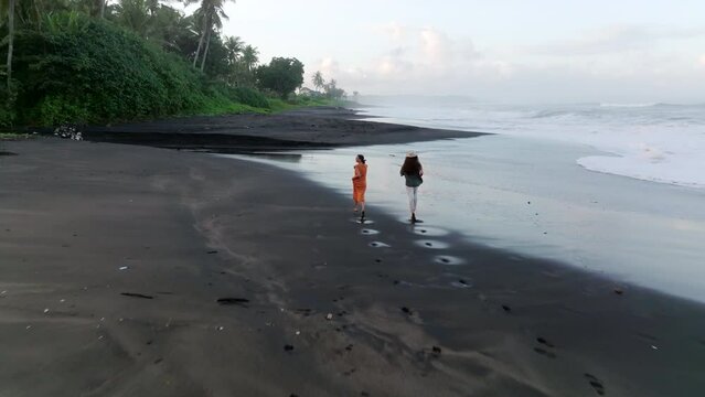 Aerial shot tracking young couple running on a black sand beach in Bali, Indonesia.