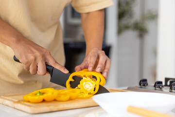 Asian man hand using knife cutting fresh yellow bell pepper on cutting board for making salad in the kitchen at home. Happy guy cooking and having dinner eating healthy food on summer holiday vacation