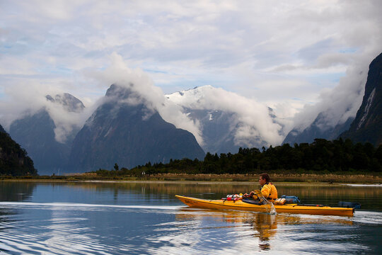 Solo kayaker paddling through calm waters of Milford Sound, surrounded by dramatic mountains and clouds.