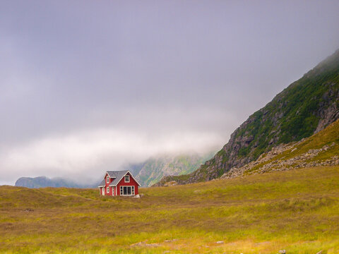 A solitary red house stands in a vast field with misty mountains in Norway, creating a peaceful rural scene.