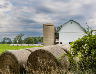 Round bales of hay in the foreground with a small white dairy barn in the background with a concrete silo and solar panels and a sky with storm clouds. © Margaret Burlingham