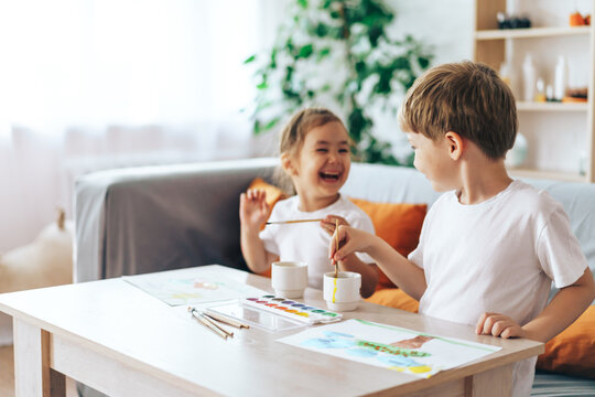 Laughing children paint pictures sitting at the table.