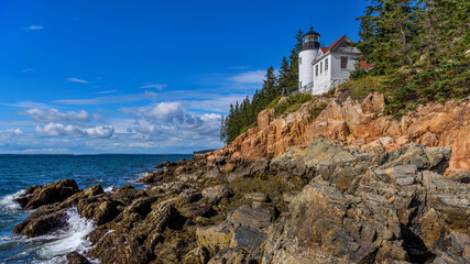 Bass Harbor Head Light - A wide-angle view of Bass Harbor Head Lighthouse standing on top of colorful seaside cliff against blue sky on a sunny Autumn morning. Acadia National Park, Maine, USA.
