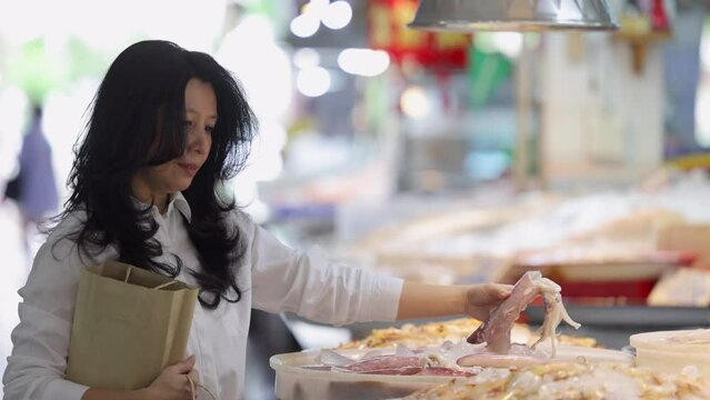 Asian woman holding paper shopping bag and pick the best fresh squid that preserved on ice for freshness from counter. Piles of fresh squid sold in seafood department of supermarkets or fresh market.