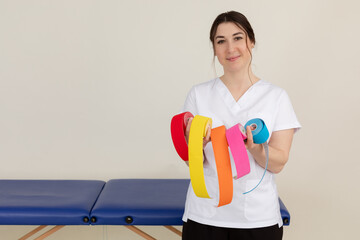 Portrait of young wonderful masseuse woman wearing white uniform, showing colourful kinesio tapes...