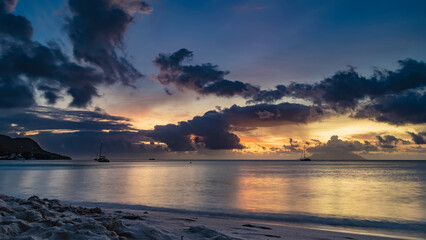 A delightful sunset over the ocean. Dark blue clouds in the sky, illuminated with golden and pink. Reflection on calm water. Footprints on a sandy beach. Long exposure. Seychelles. Mahe. Beau Vallon