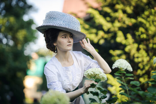 Portrait Of A Young Slender Woman In A 1910s Costume. A Lady In A Hat In The Fashion Of The Early 20th Century Walks In A Garden