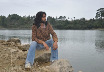 A good looking young man with long hair and beard looking sideways while sitting on stone by the lake