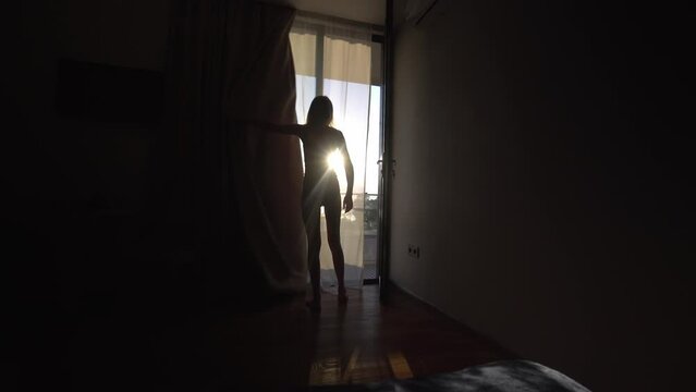 Woman in panties opens curtains after sleeping in luxurious room at Bali Hotel overlooking terrace and sun. Woman woke up and stood in front of window. Girl opens curtains and meets sunrise