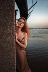 A beautiful slender girl with red hair and a perfect figure stands in the river at sunrise in a pink and gold satin tight-fitting sundress with lowered straps. Gentle sexuality, romance and femininity