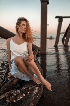A beautiful slender barefoot ginger girl with a perfect figure in a skirt and a T-shirt of delicate colors is sitting on a log sticking out of the river. Sensual femininity and romantic image