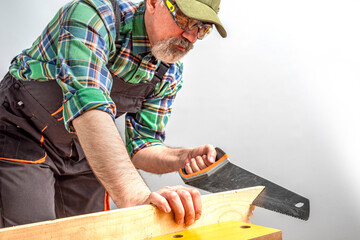 Aged worker sawing a board with a saw using vise sawhorse. Senior man in overalls, cap and goggles