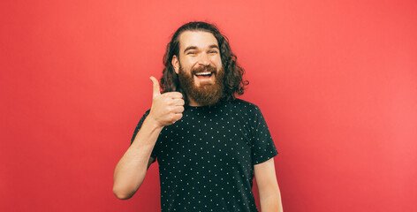 Portrait of a bearded man with long curly hair standing near a red background wearing a black...