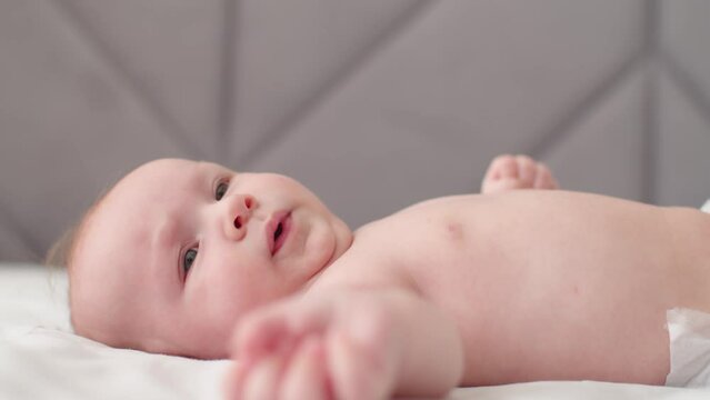 Beautiful newborn baby., closeup face. Cute tiny baby with bare belly looks up, slow motion. Active newborn boy lies on the bed and moves his arms. Low angle view of a chubby male baby..