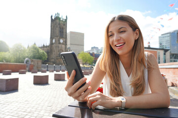Smiling girl with mobile phone in her hands sits at the table outdoors with the cityscape on the background. Beautiful young woman is watching videos and photos on her smartphone at the cafe table. - obrazy, fototapety, plakaty