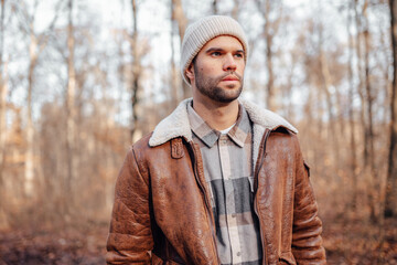 A portrait of a young white man posing in the woods dressed in autumn clothes: brown jacket,...