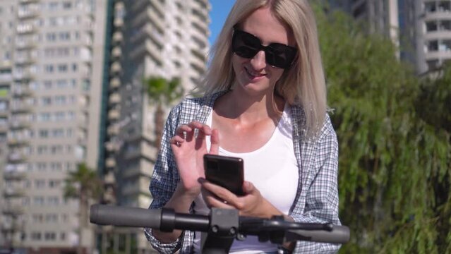 Blonde woman with black glasses uses app on her smartphone to unlock shared electric scooter on city street. New eco-friendly transport. Woman scans QR code with smartphone