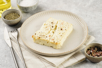 oven baked sheep cheese feta with herbs, oil and spices on round off-white plate, kitchen napkins, grey background