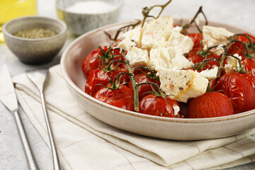 Roasted red tomatoes on branches with seasoned feta cheese on round off-white plate on kitchen napkins on grey background