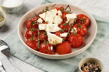 Roasted red tomatoes on branches with seasoned feta cheese on round off-white plate on kitchen napkins on grey background