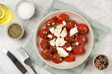Roasted red tomatoes on branches with seasoned feta cheese on round off-white plate on kitchen napkins on grey background