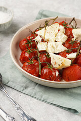 Roasted red tomatoes on branches with seasoned feta cheese on round off-white plate on kitchen napkins on grey background