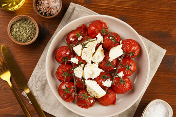 Roasted red tomatoes on branches with seasoned feta cheese on round off-white plate on kitchen napkins on wooden background