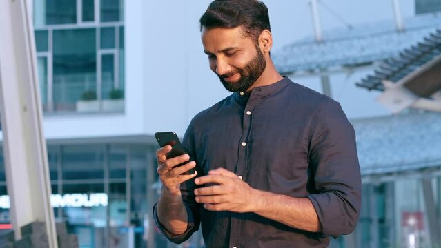 Smiling indian business man, eastern professional businessman holding cellphone tech using mobile phone banking app, making e payment, texting message standing in urban city on modern street outdoors.