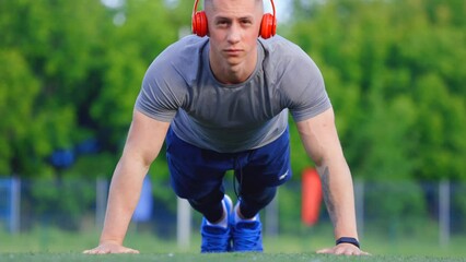 Muscular sportsman does push-ups while listening to music with headphones in an outdoor stadium. Summer training of a serious Caucasian athlete at the stadium before the competition