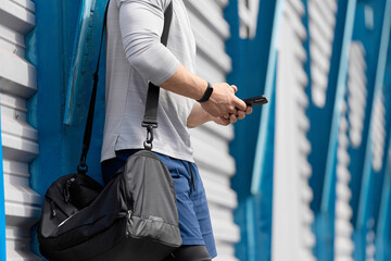 Male athlete with black bag on his shoulder and smartphone in his hands stands near stadium waiting for training. strong man with a sports bag came to the stadium, holding a cell phone in his hands