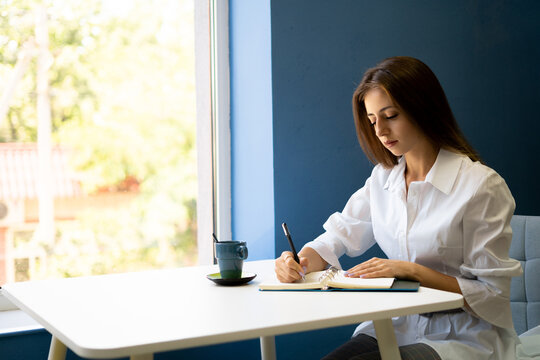 Serious female student writes a story in a notebook while sitting in a cafe with a cup of cappuccino. Focused young woman notes in notepad