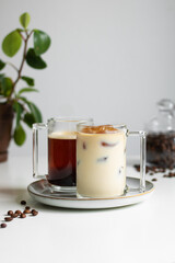 Two cups of coffee. Hot coffee in the background, coffee ice with milk in the foreground (a coffee drink made from ice cubes of coffee). Behind the coffee cups is a jar of coffee beans and a flower. 