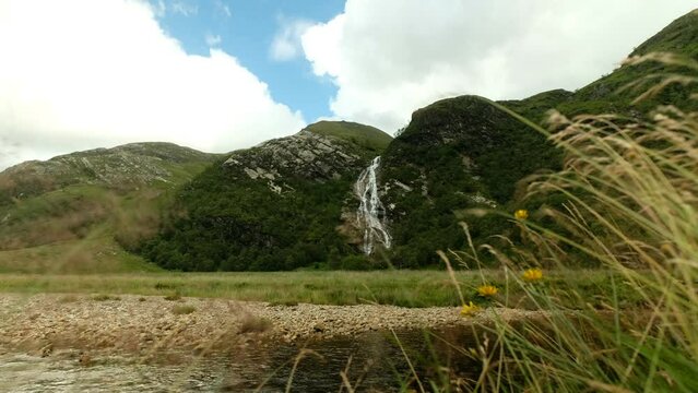 A tall distant waterfall, runs calmly along the mountain creek, as seen through some green grass in the foreground in a windy summer day. a 4K video clip. Ben Nevis, Scotland.