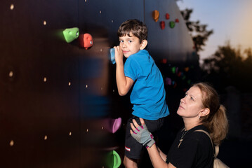 Little boy doing sports with mom in the evening, boy climbs the rock climbing wall, mom holds him...