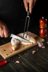The chef or cook cuts a fresh raw chicken drumstick on a dark background. Nearby on the kitchen table are ingredients for cooking and spices