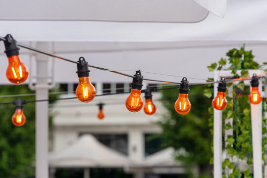 Light garland on the street at daylight. Decoration of the street with vintage Edison bulbs garland, festive city decorations for cafe and restaurant, celebrating, lights as exterior decor