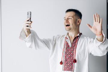 Guy in Ukrainian vyshyvanka stands holding phone takes selfie and smiles.