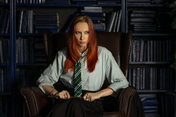 A young red-haired witch with a magic wand and a haughty expression sitting in an old library,...