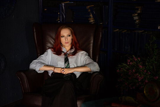 A young red-haired woman with a haughty expression on face sitting in an old library in leather chair. She is dressed in a white shirt, black skirt and green tie. Mafia princess, librarian or whitch.