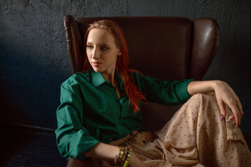 A beautiful natural red-haired girl in a green shirt and a long linen skirt, in a vintage interior, poses sitting in an old vintage leather armchair, the rays of the sun fall on her. Hippie