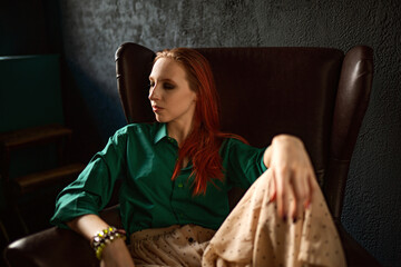 A beautiful natural red-haired girl in a green shirt and a long linen skirt, in a vintage interior, poses sitting in an old vintage leather armchair, the rays of the sun fall on her. Hippie