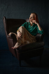 A beautiful natural red-haired girl in a green shirt and a long linen skirt, in a vintage interior, poses sitting in an old vintage leather armchair, the rays of the sun fall on her. Hippie