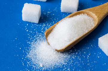 Heap of white granulated sugar in spoon with sugar cube on blue background