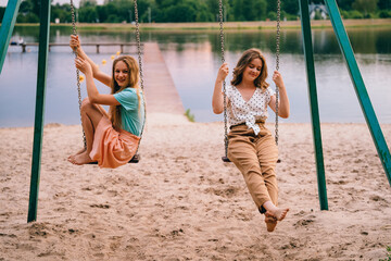 Two young women playing with swing over lake at sunset. Teenagers having friends fun together while talking and swinging