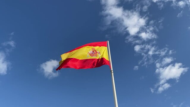 Flag of the Kingdom of Spain waving in the wind on a mast; red, yellow, shield and crown, blue sky in the background; country of European Union, also symbolizes Spanish language and Latin countries