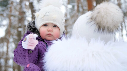 on the background of the winter forest, a portrait of a small one-year-old girl in a funny hat with a bumbon. the girl reaches into the camera with her hand. High quality photo
