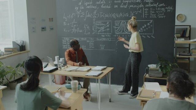 Tilt down shot of college girl standing by chalkboard in classroom and giving presentation in front of diverse group mates and African American professor