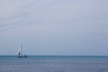 A sailing yacht moves on the surface of a calm sea at the horizon.