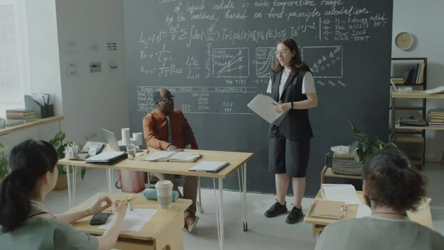 Tilt down shot of female student holding papers and giving presentation in front of group mates and African American professor during university class